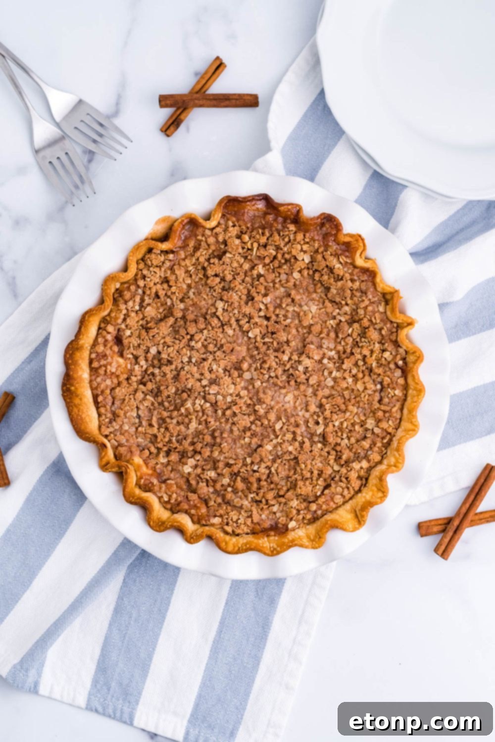 Apple Crumble Cheesecake Pie on table with cinnamon sticks and towel by pan 