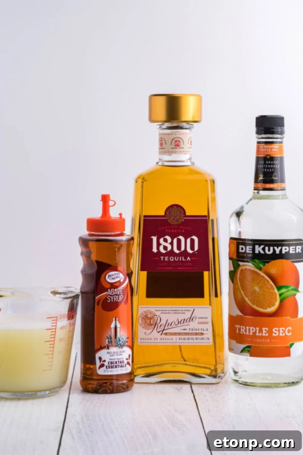 Fresh ingredients for making a batch of margaritas, including limes, lemons, and bottles of spirits, arranged on a rustic wooden table