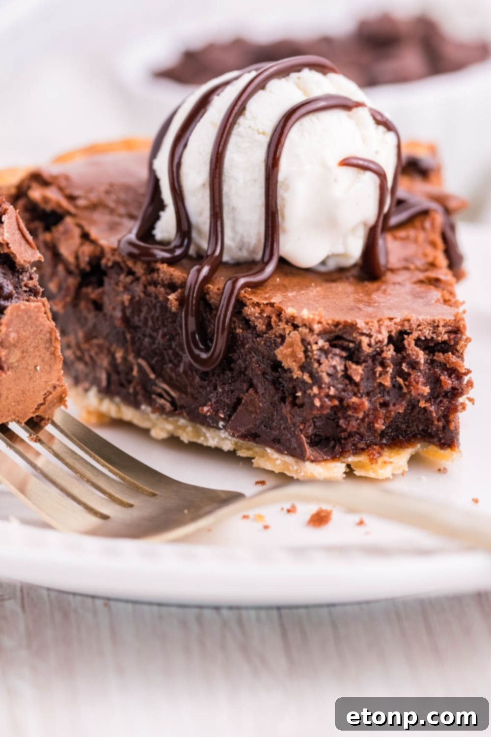 A slice of brownie pie on a plate, topped with hot fudge and a scoop of vanilla ice cream, melting slightly.