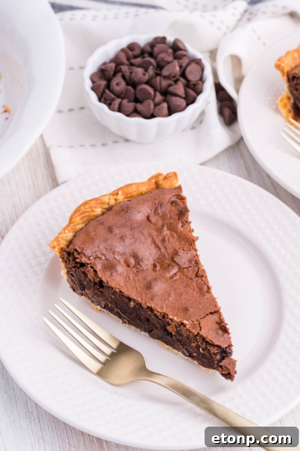 A chocolate pie on a white plate with a bowl of chocolate chips blurred in the background, creating an inviting scene.