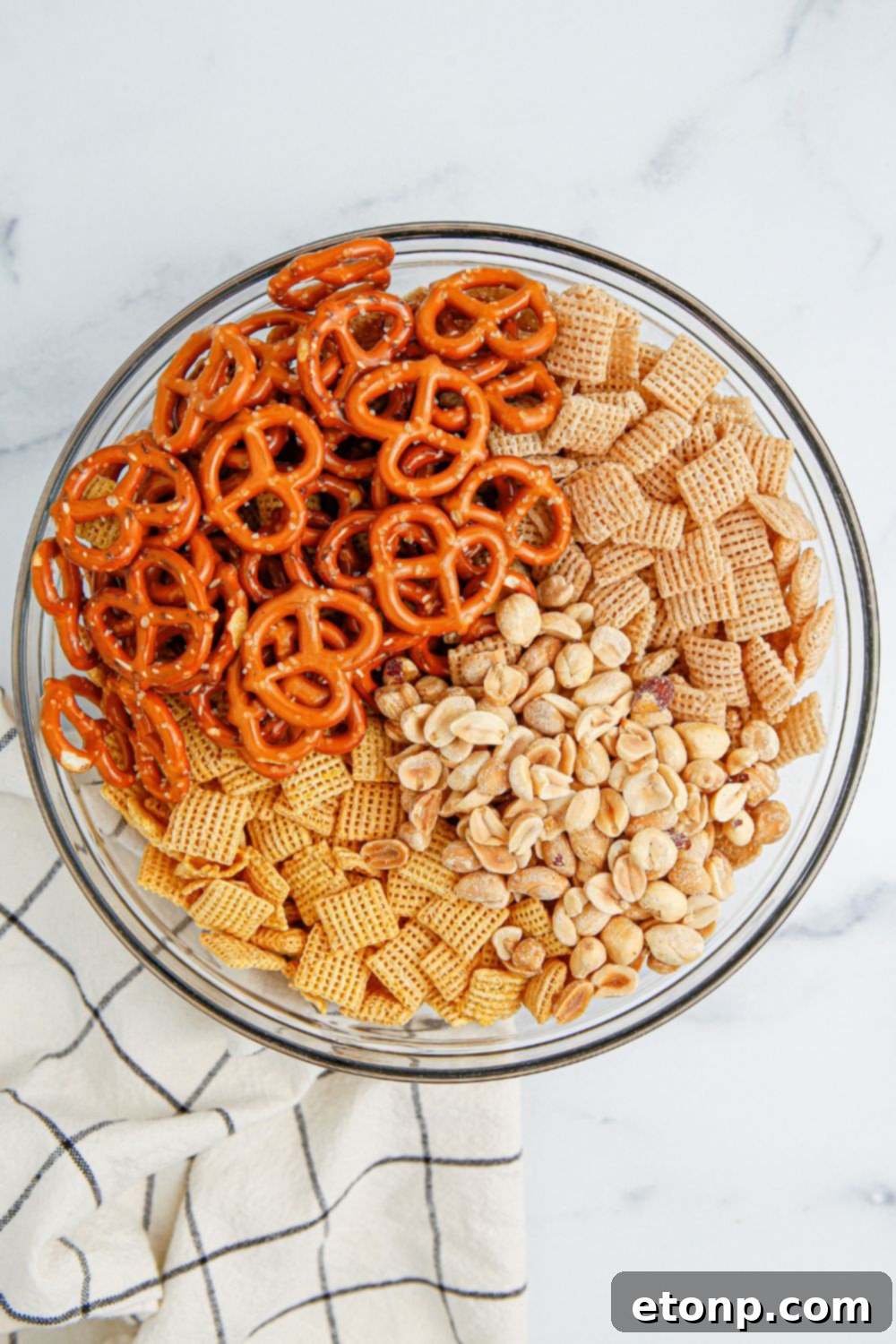 A glass bowl showcasing the mixed dry ingredients for the snack mix, including cereals, pretzels, and peanuts.