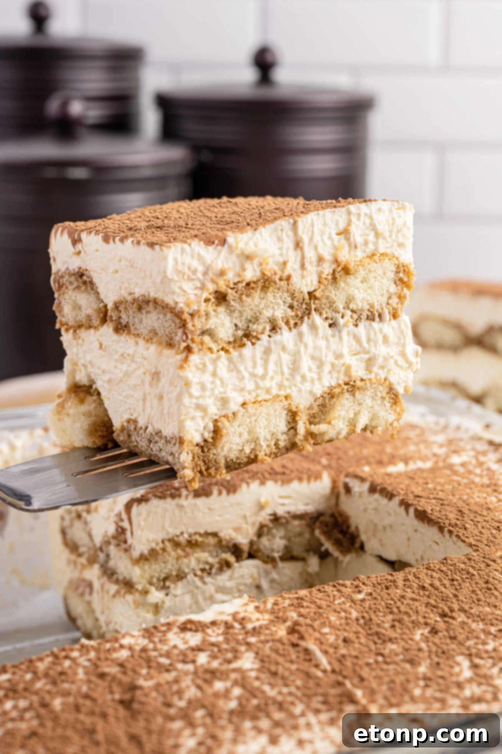 A perfectly portioned slice of tiramisu being gently lifted from a baking dish with a spatula, revealing its beautiful layers of coffee-soaked ladyfingers and creamy mascarpone filling, dusted with cocoa powder.