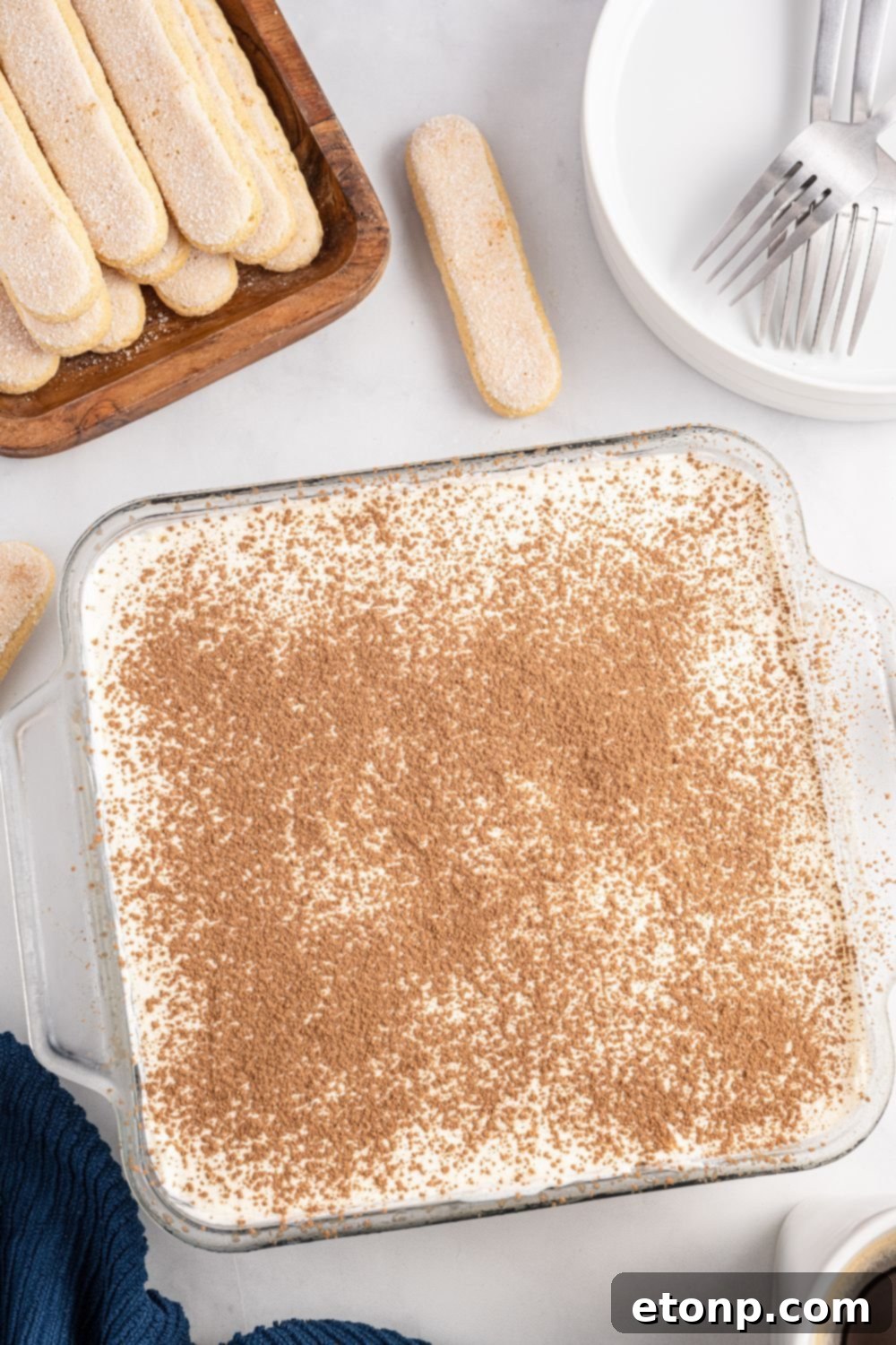 An inviting overhead view of a partially assembled tiramisu in a clear glass pan, sitting on a table, with layers of ladyfingers and creamy filling visible.