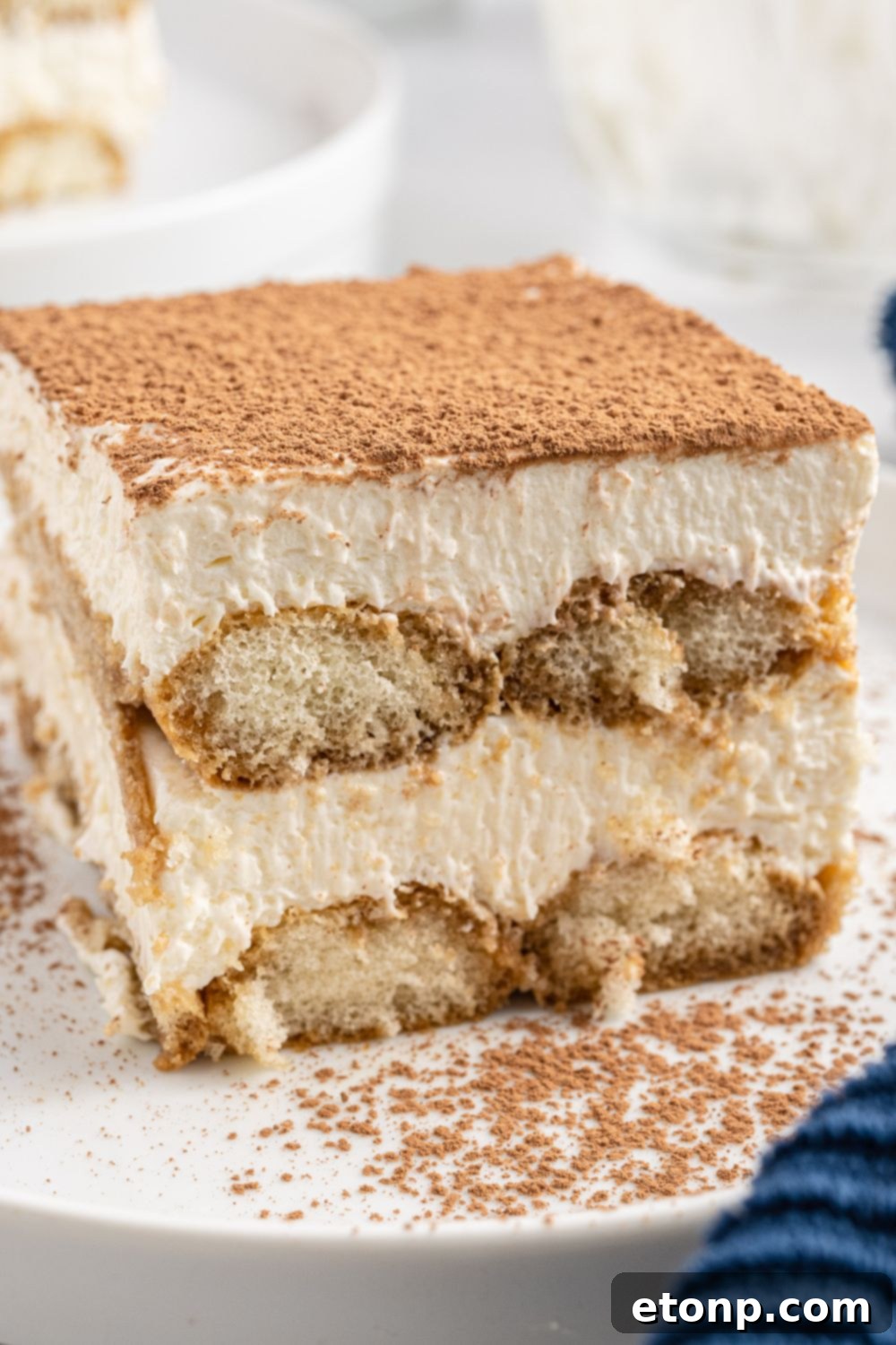 A perfectly sliced piece of tiramisu presented on a white plate, generously dusted with cocoa powder on top, ready to be enjoyed.
