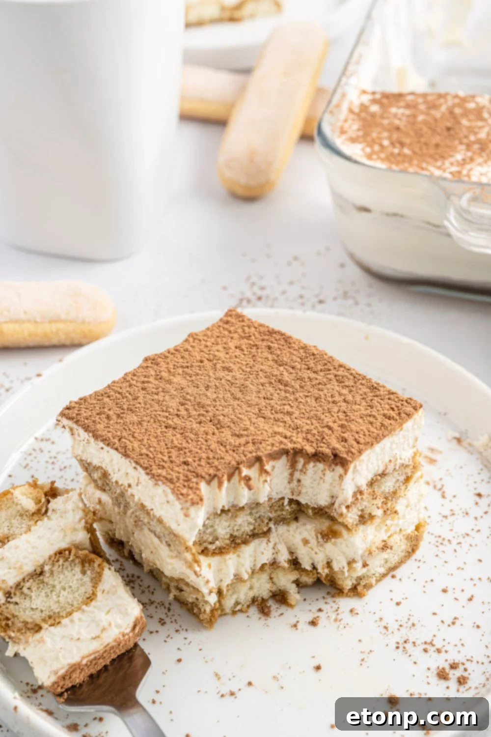 A beautifully presented slice of tiramisu cake on a white plate, with a fork gently placed beside it, ready to be enjoyed.