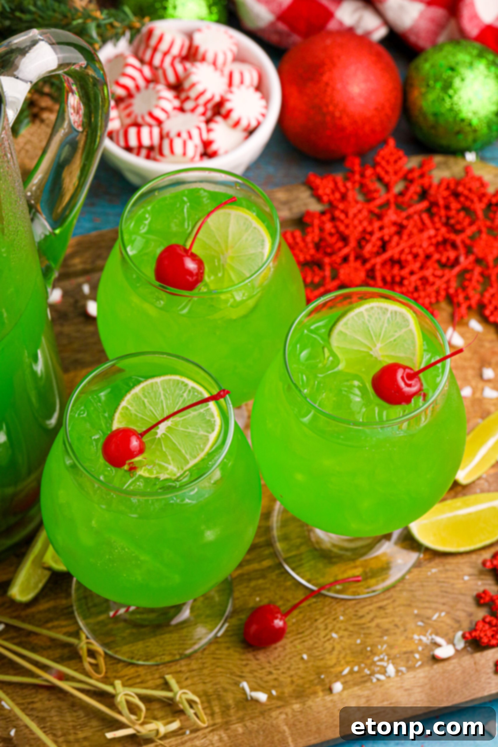 A glass of Grinch Punch, garnished with a cherry and a lime wedge, placed on a table with a bowl of peppermints in the background, creating a festive scene.