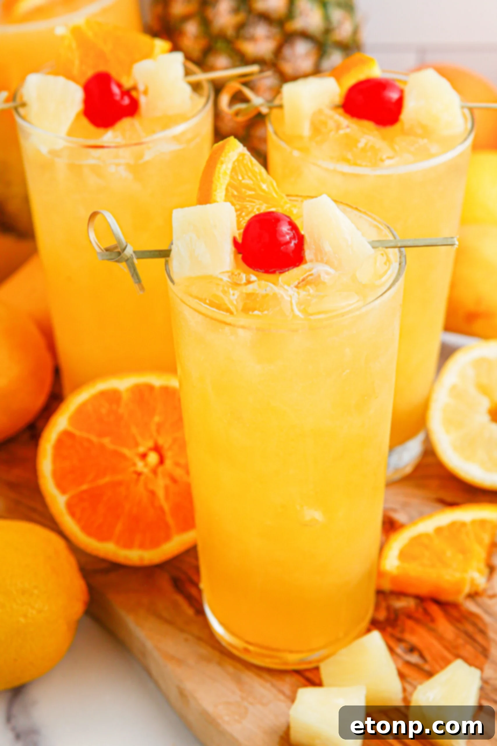 Tropical Lemonade Punch in 3 tall clear glasses on a wooden table with fruit around it and a pineapple in the background