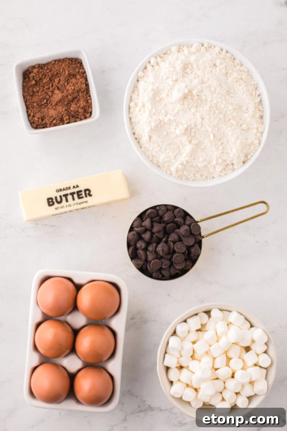 Ingredients for hot chocolate cookies, including sugar cookie mix, chocolate chips, cocoa powder, and marshmallows, laid out on a table.