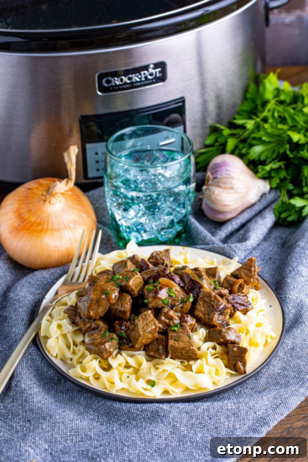 staged photo of crockpot in back, onions and garlic, and a plate with steak bites on it and a fork on side 