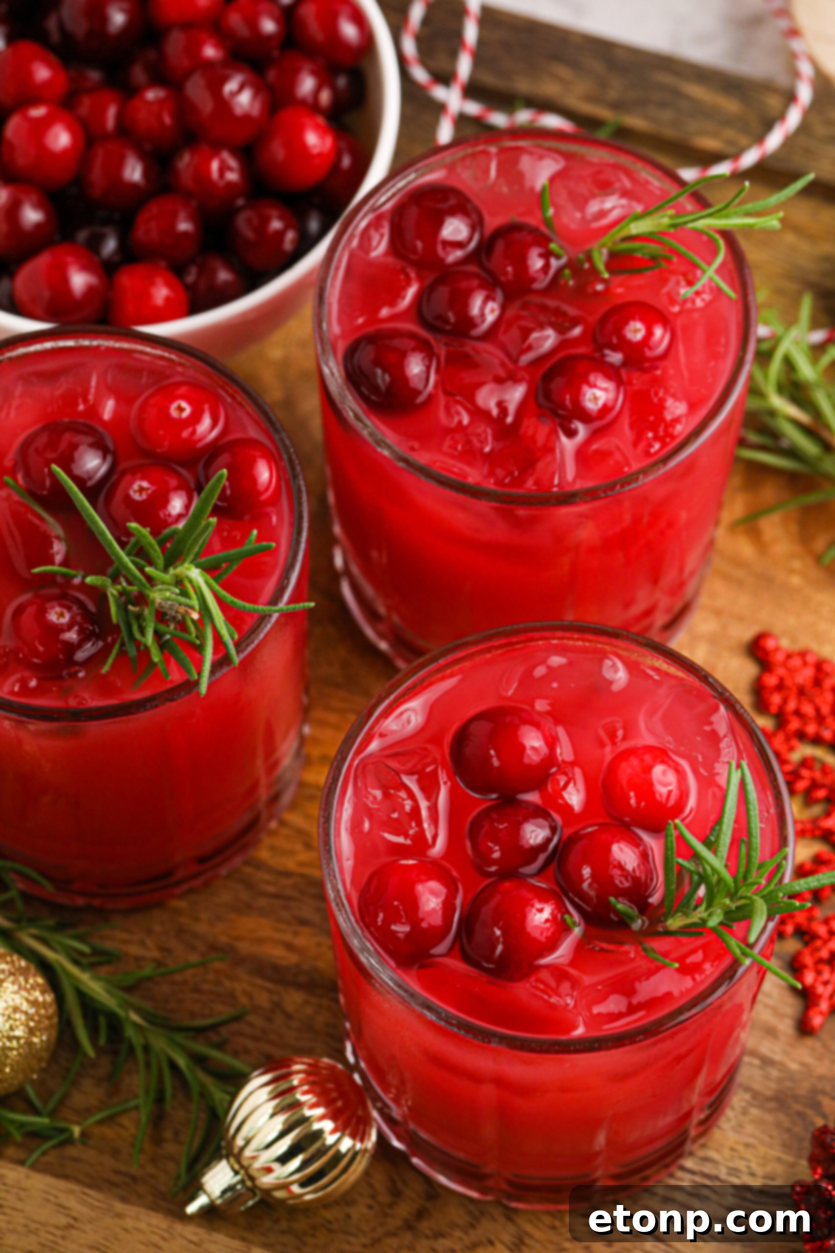Three elegant glasses of cranberry orange punch, garnished with fresh cranberries and rosemary, displayed on a rustic wooden board.