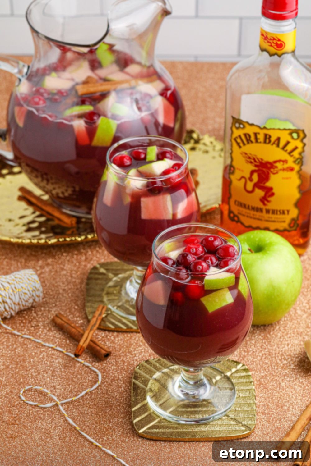 Cinnamon Spice Sangria 2 Glasses filled with Fireball Sangria, featuring rich red wine, cinnamon whiskey, and slices of apple and cranberry, with a bottle of whiskey in the background.