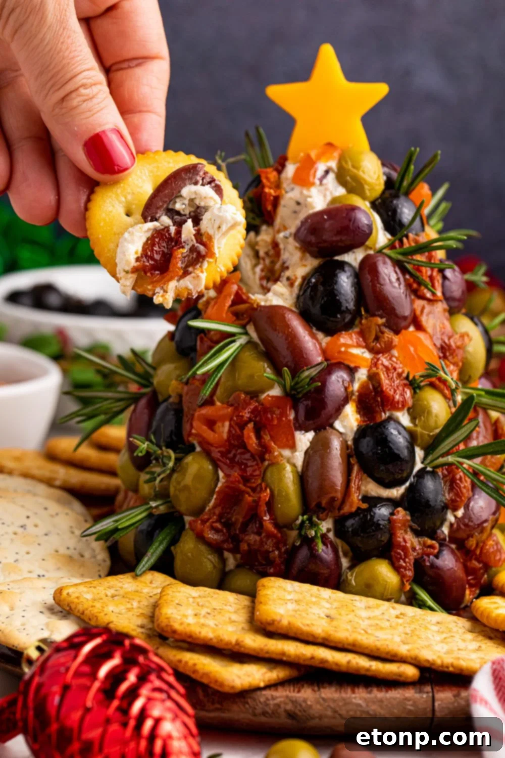 A cracker artfully topped with cream cheese and an olive dip, positioned next to the festive cheeseball on the table, inviting guests to dig in.