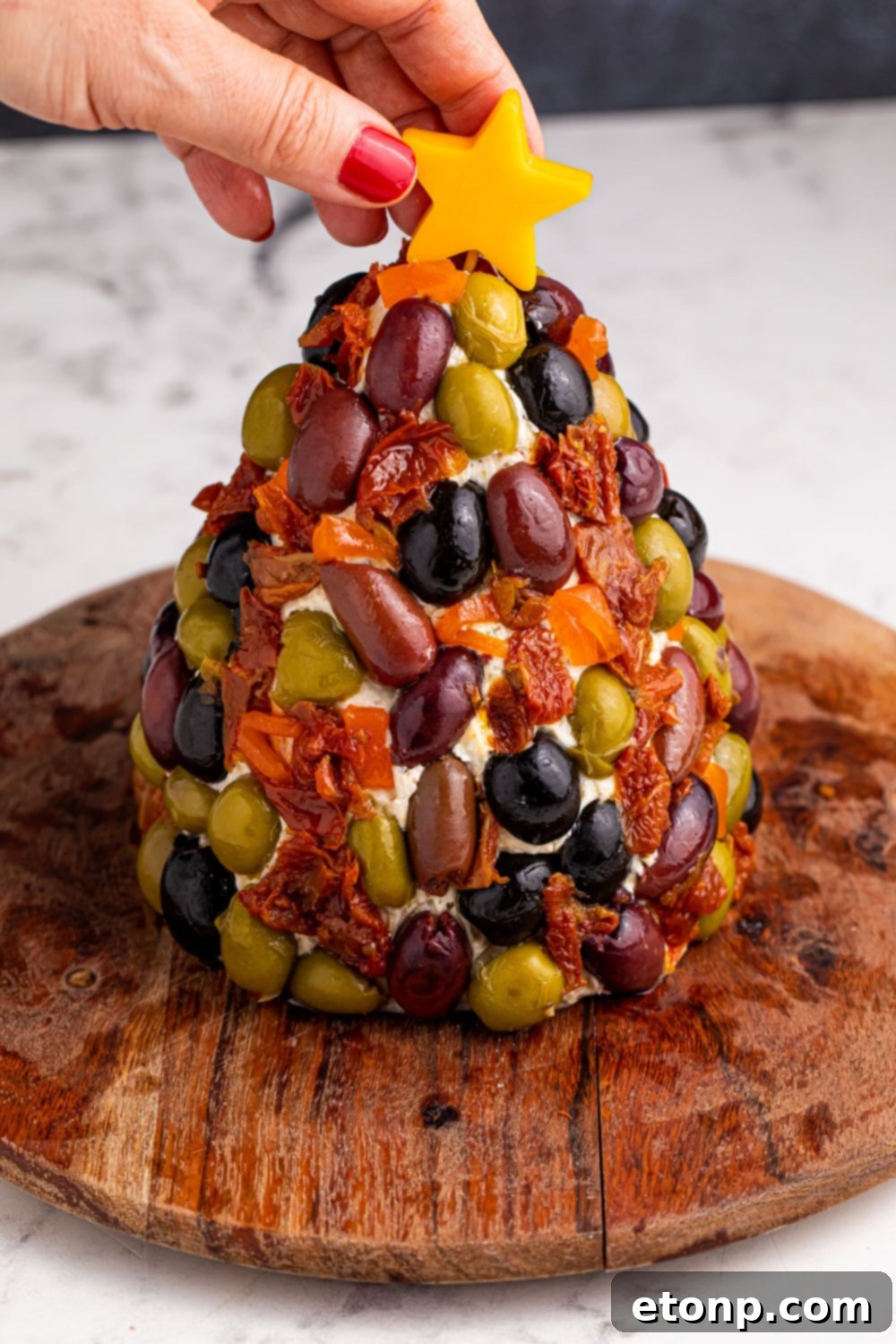 A close-up of the fully decorated holiday cheeseball, with a bright cheese star being gently placed on top as the final festive touch.