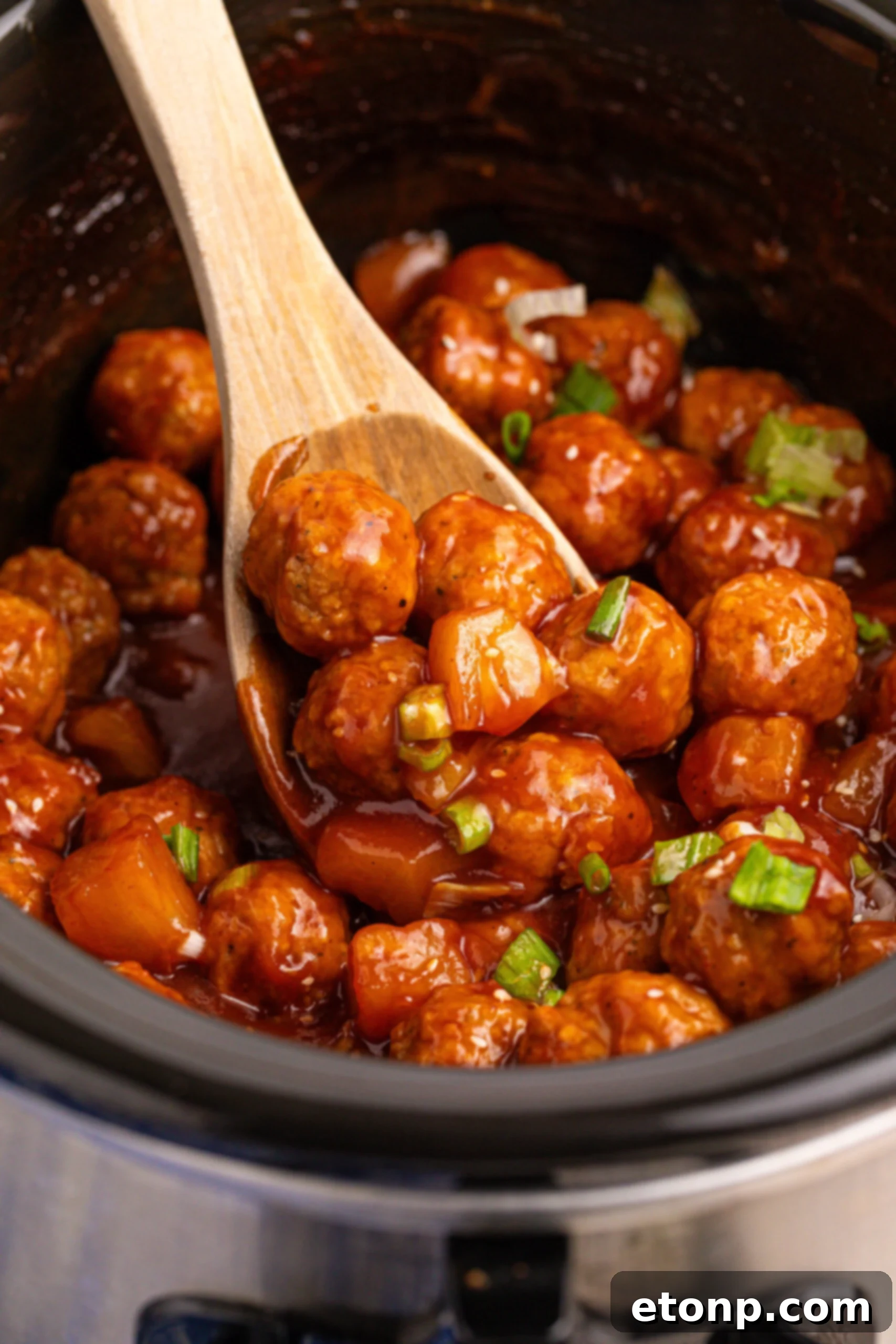 Slow Cooker 3 Ingredient Hawaiian Meatballs in a black crock pot with a wooden spoon lifting up a few 