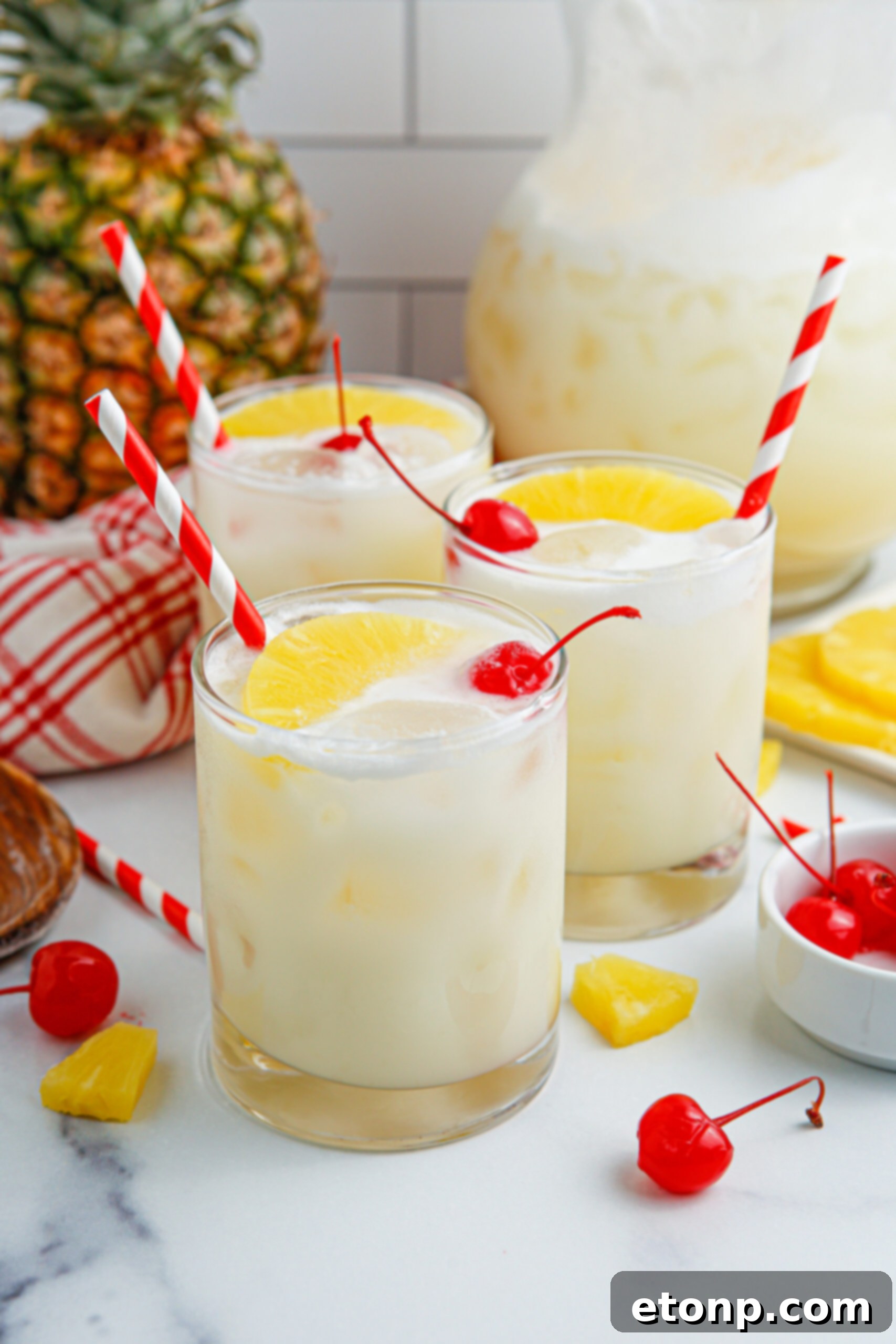 Close-up shots of three Pina Colada Punches in clear glasses sitting on a table, garnished with pineapple wedges and cherries.