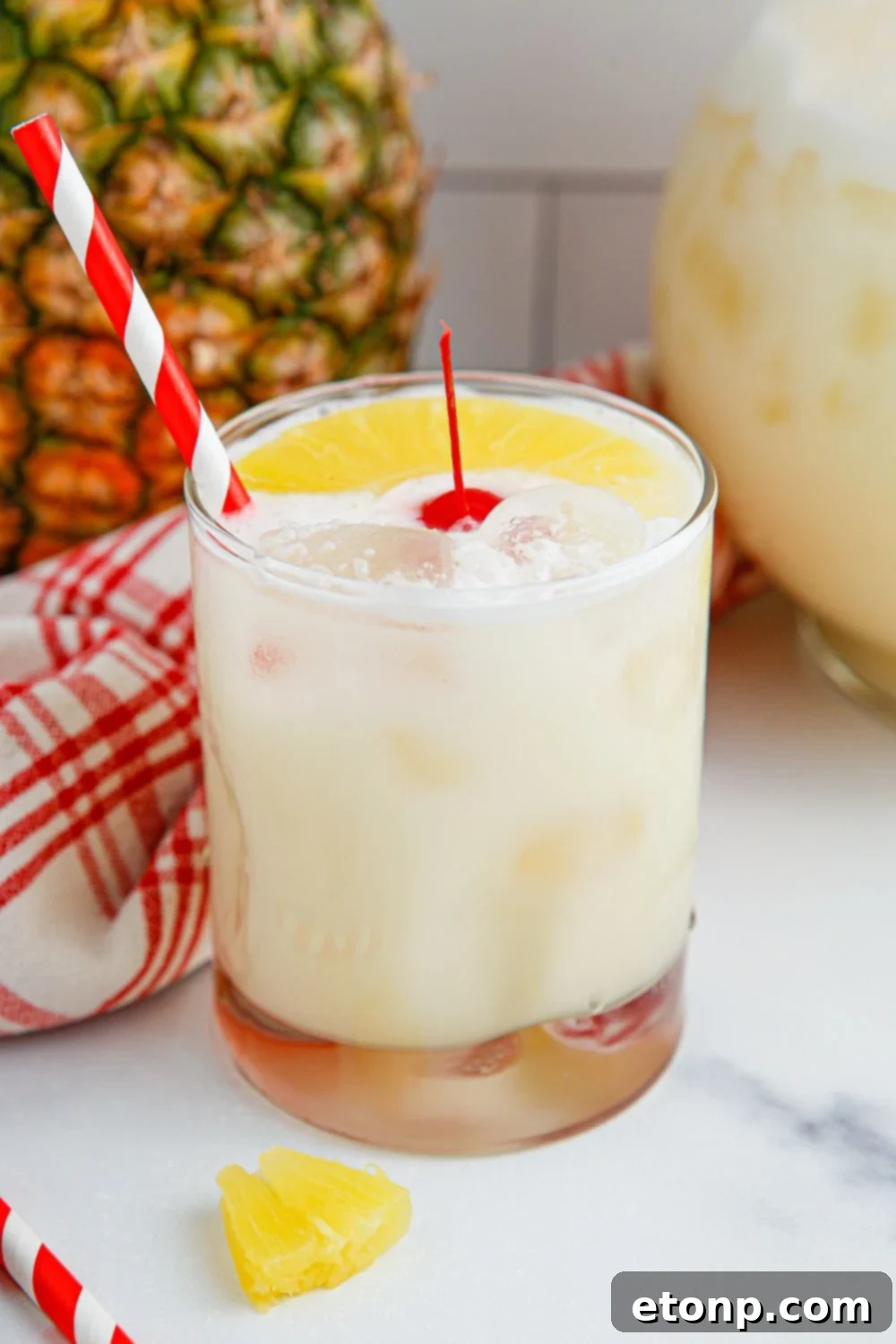 Pina Colada Punch sitting on a table in a garnished glass, with a pitcher of punch and fresh pineapple in the background.