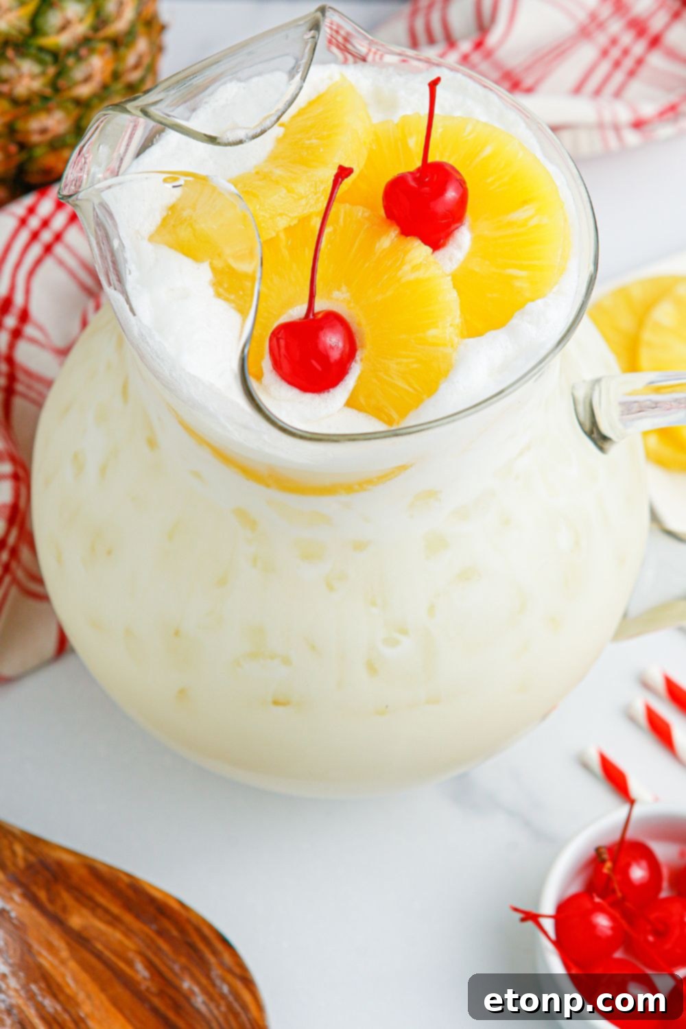Pina Colada Punch in a pitcher with cherries and pineapple slices on the table, alongside a wooden spoon.