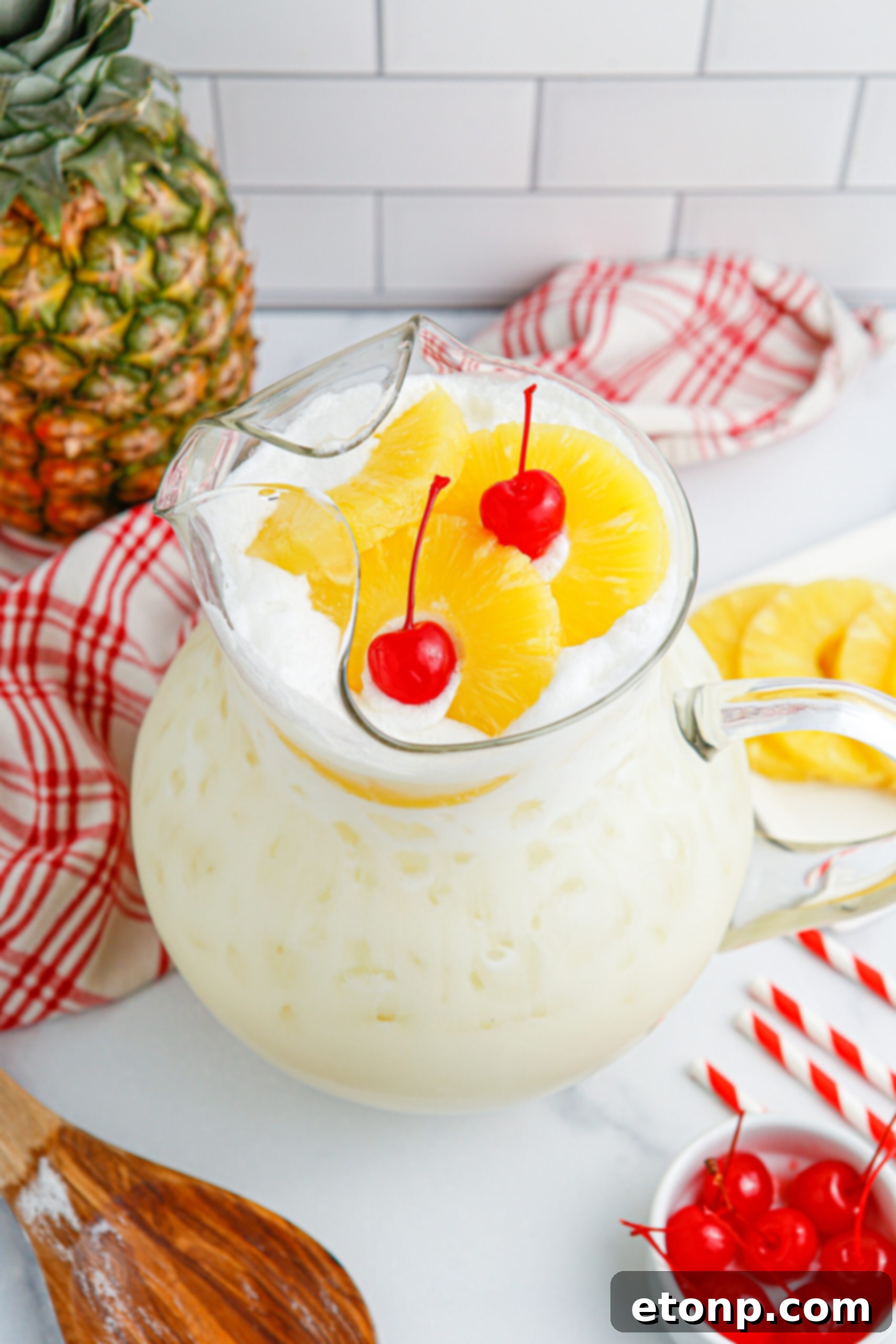 Pina Colada Punch in a pitcher with cherries and pineapple slices on the table, alongside a wooden spoon.