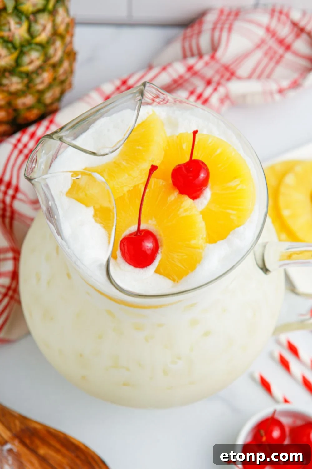 Pina Colada Punch in a pitcher on a table, beautifully garnished with fresh pineapple slices and maraschino cherries.