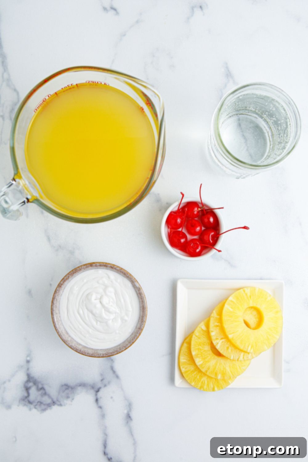 Pina Colada Punch ingredients displayed on a table: a pitcher of pineapple juice, a can of cream of coconut, sparkling water, maraschino cherries, and fresh pineapple wedges.