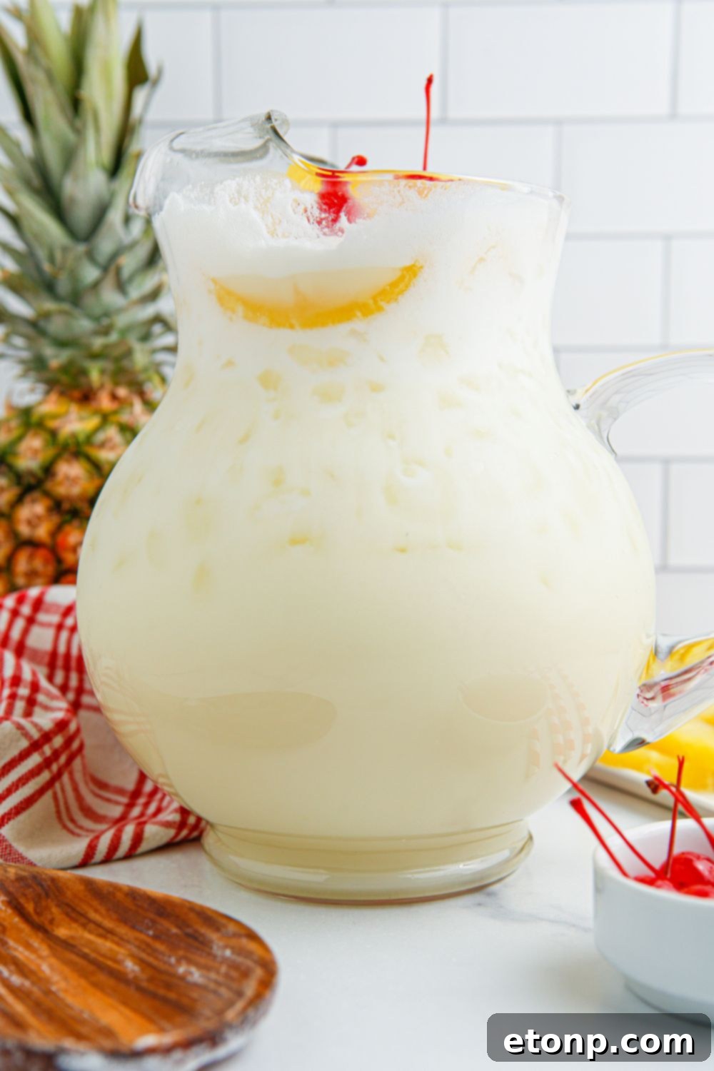 Pina Colada Punch in a pitcher, surrounded by a whole pineapple and a bowl of bright red cherries, ready for garnish.