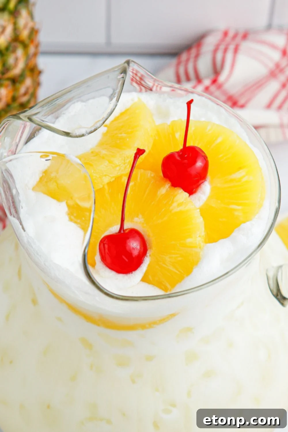 Close-up of Pina Colada Punch in a pitcher, featuring pineapple slices and cherries floating beautifully on top.