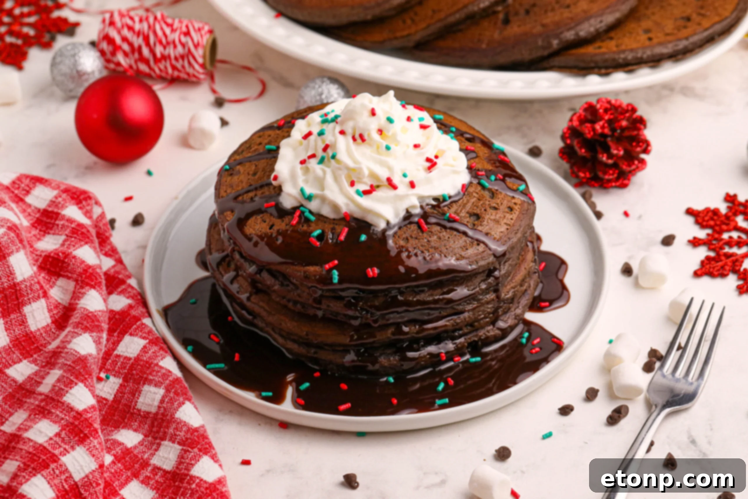 An inviting overhead shot of Hot Chocolate Pancakes, beautifully stacked on a white plate, generously topped with syrup, whipped cream, and festive holiday sprinkles.