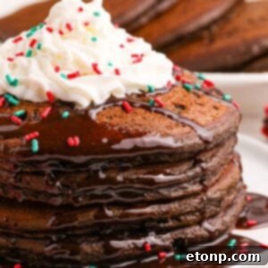 Hot Chocolate Pancakes stacked on a plate topped with syrup and whipped cream and holiday sprinkles