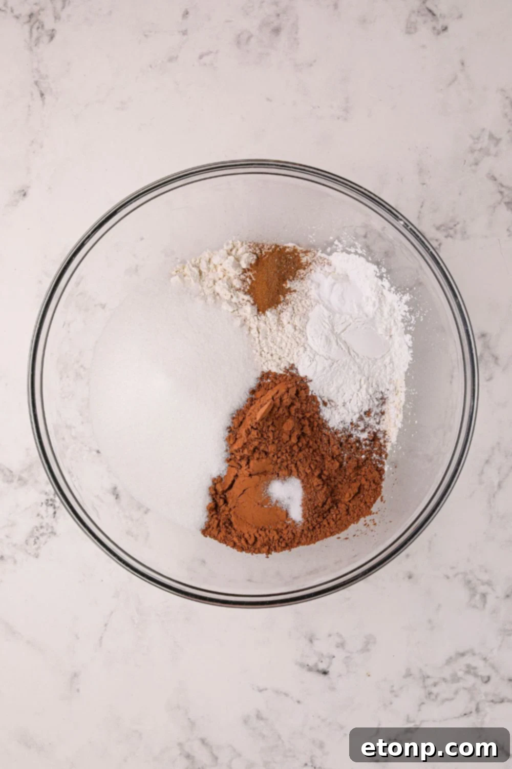 Dry ingredients for Hot Chocolate Pancakes, including flour, cocoa powder, and sugar, mixed in a clear glass bowl on a table, ready for wet ingredients.