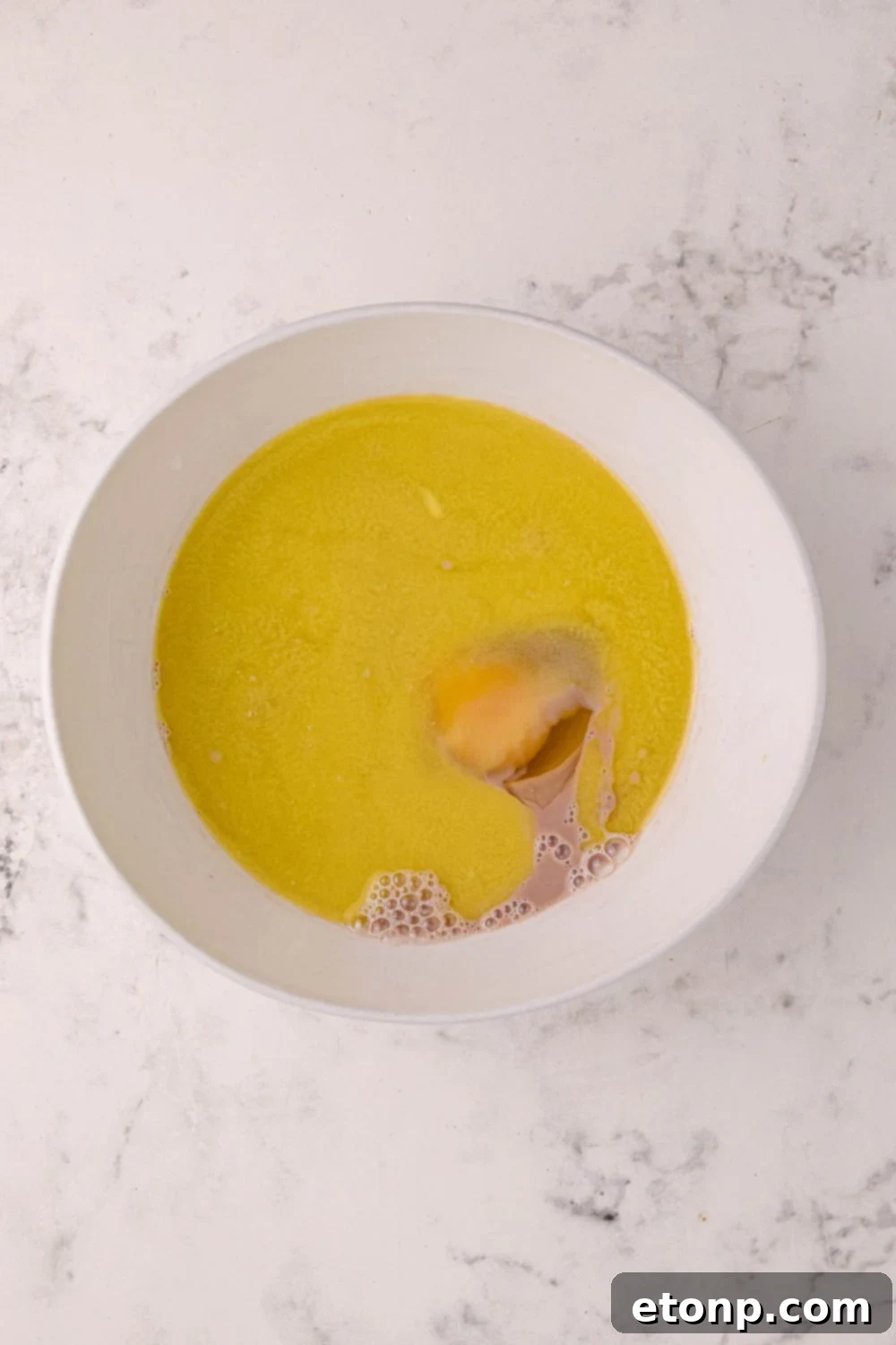 A mixing bowl on a table containing melted butter and a cracked egg, prepared to be combined with other ingredients for Hot Chocolate Pancakes.