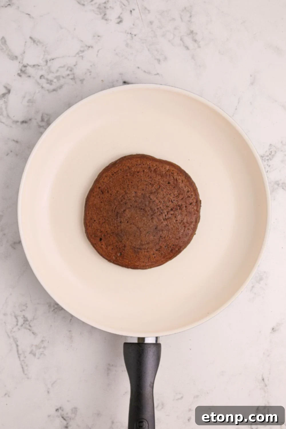 A Hot Chocolate Pancake cooking to golden perfection in a skillet on the stovetop, with small bubbles forming on its surface.