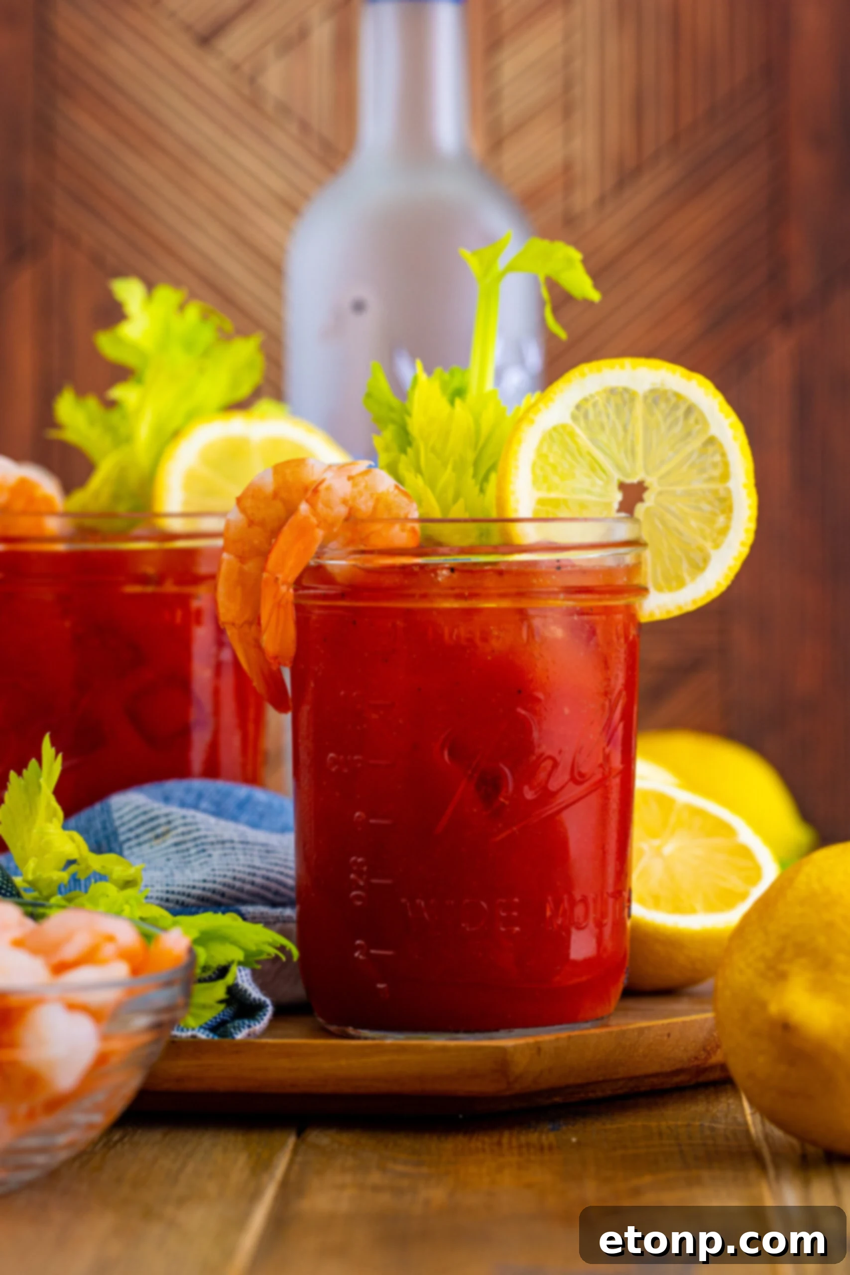 Two Bloody Mary cocktails on a table with a glass vodka bottle in the softly blurred background. The drinks are garnished with celery and lime wedges, emphasizing fresh ingredients.