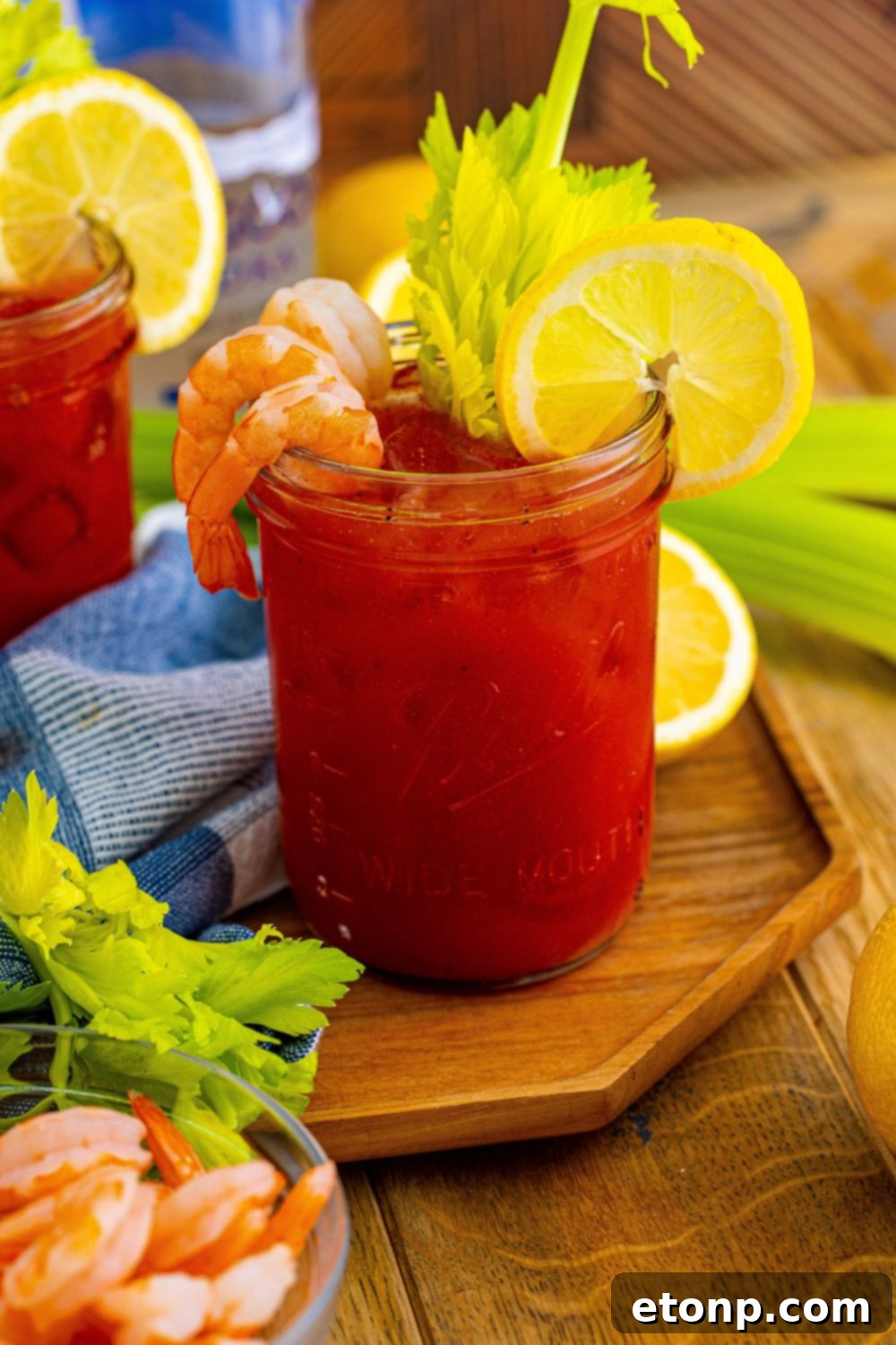 An extravagantly garnished Bloody Mary cocktail, featuring a large shrimp, lemon wedge, and celery, with various appetizers artfully arranged around the drink on a rustic cutting board.