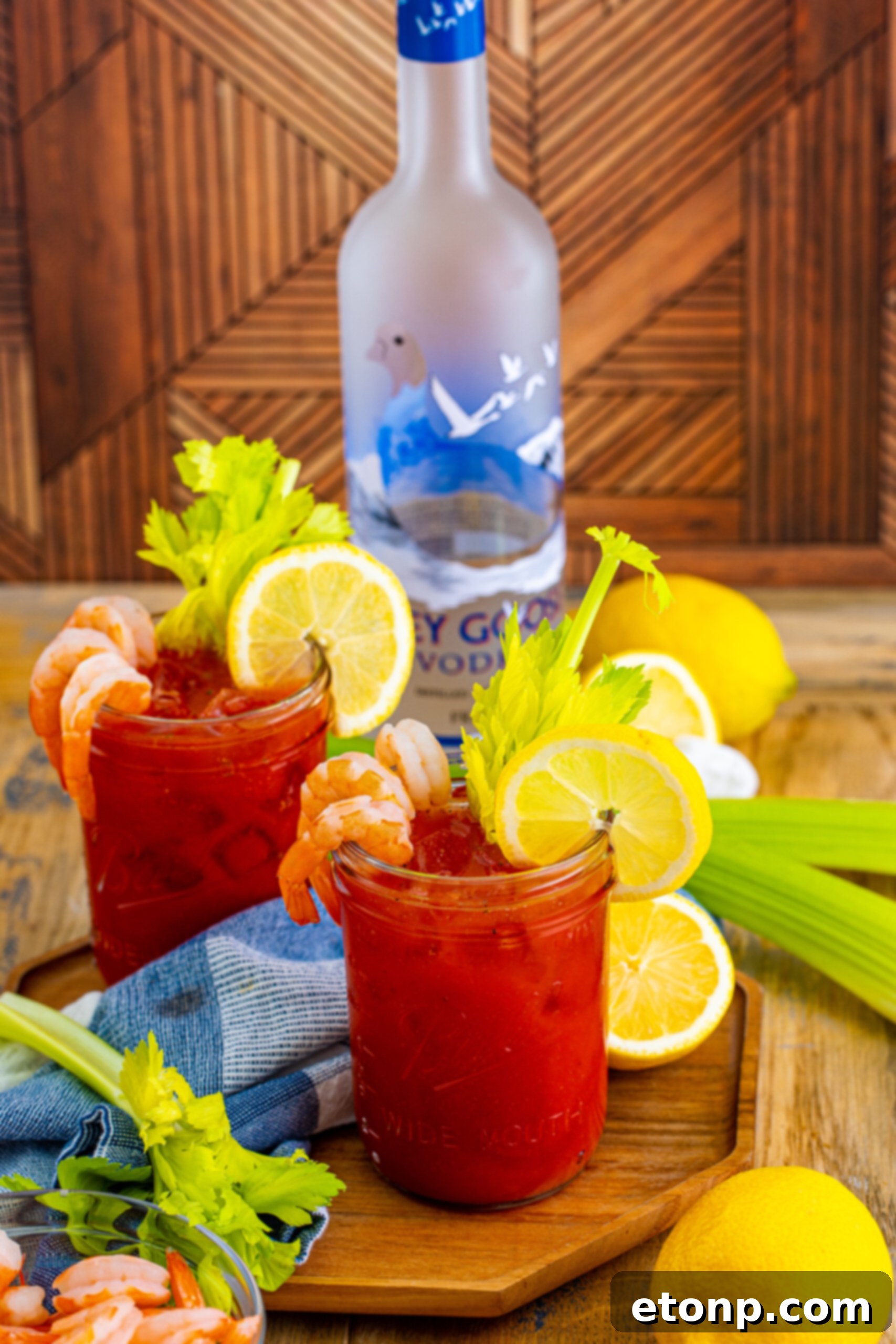 A pair of beautifully prepared Bloody Mary cocktails in glasses, with a vodka bottle subtly blurred in the background, set on a dark surface.