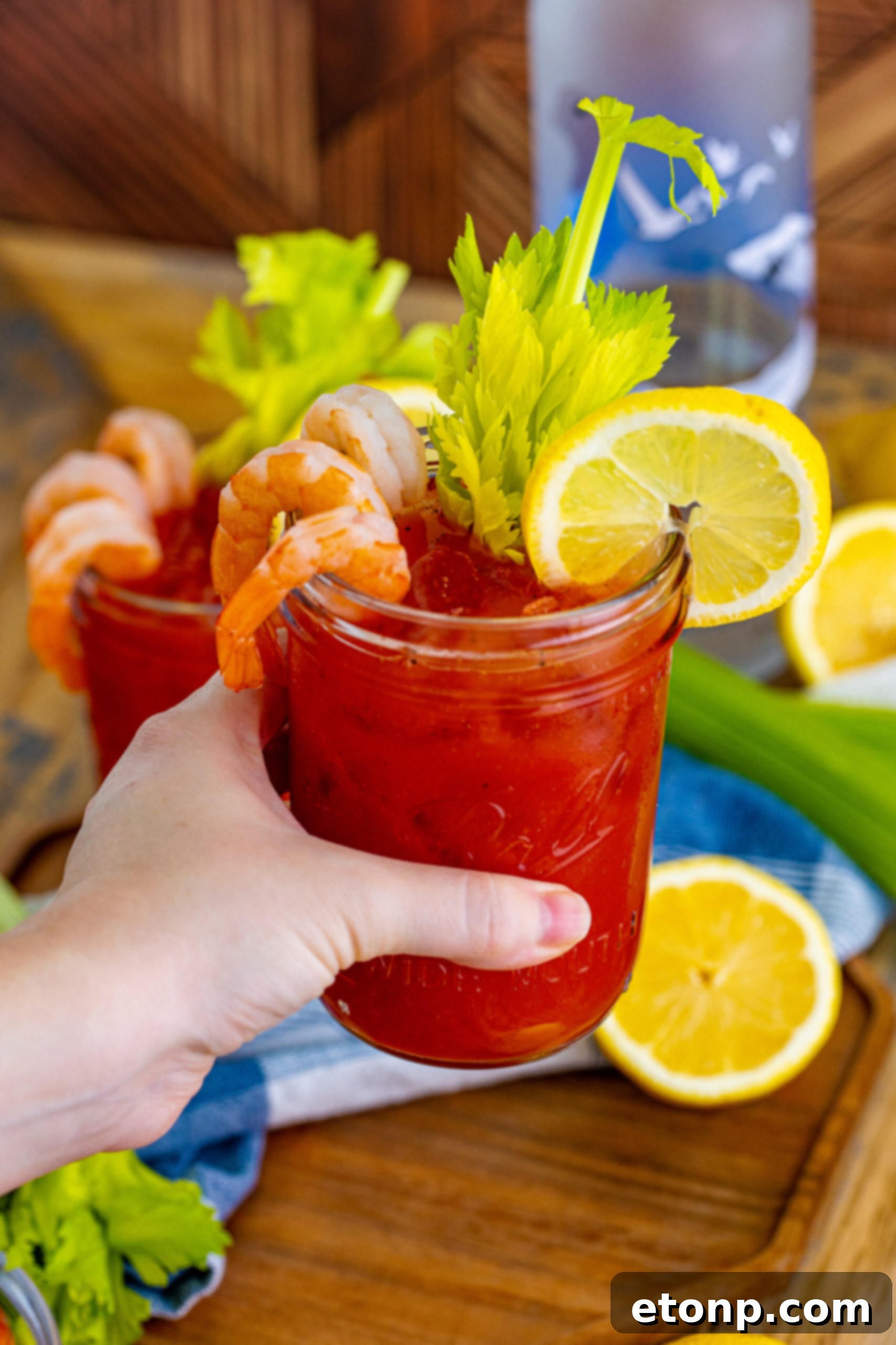 A hand holding up a Bloody Mary in a clear mason jar glass, garnished with a vibrant lemon slice and a large shrimp, against a bright background.