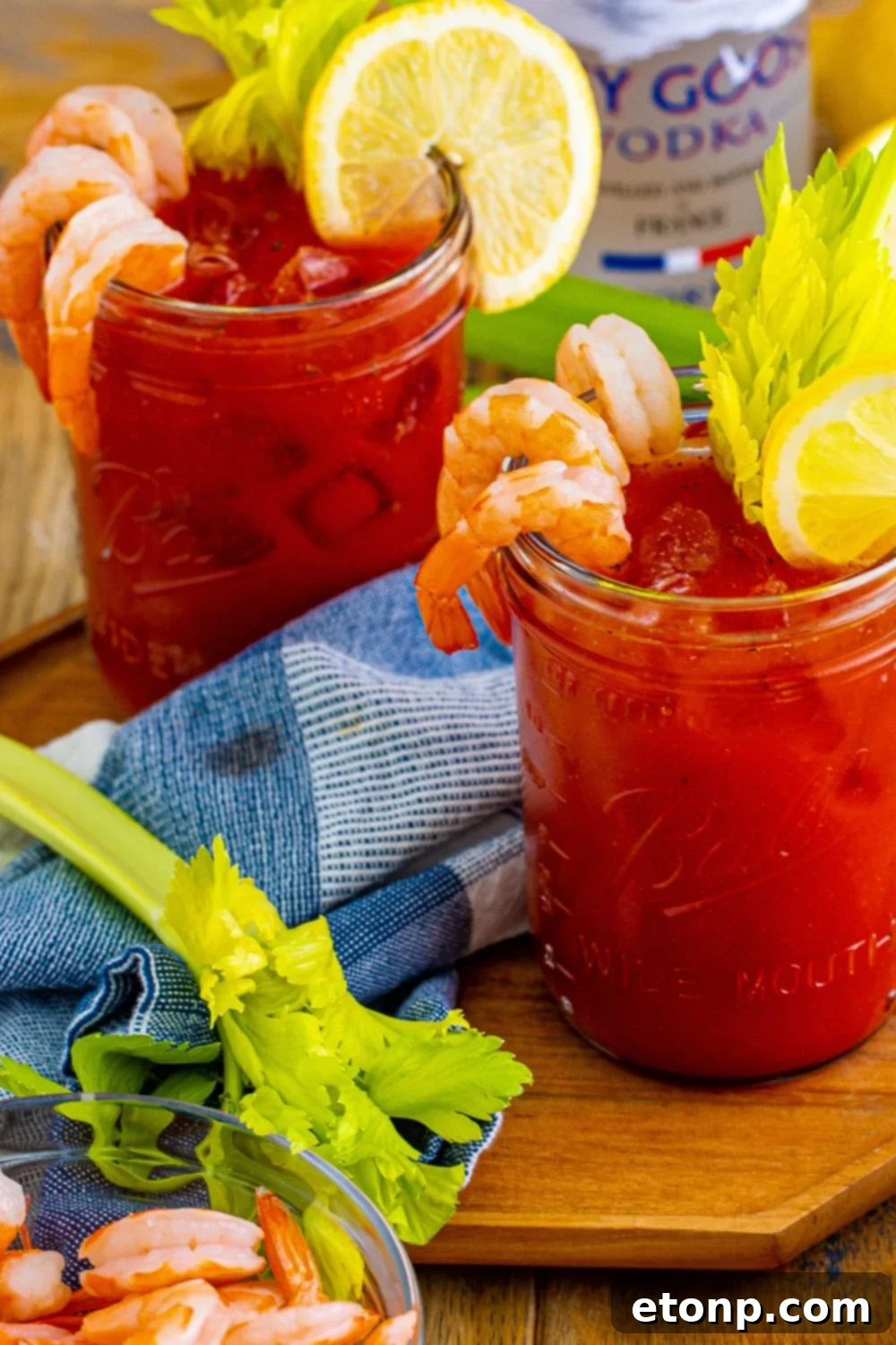 Three perfectly garnished Bloody Mary cocktails displayed on a rustic wooden table. Each glass features shrimp, a lime wedge, and a crisp celery stalk, showcasing impressive presentation.
