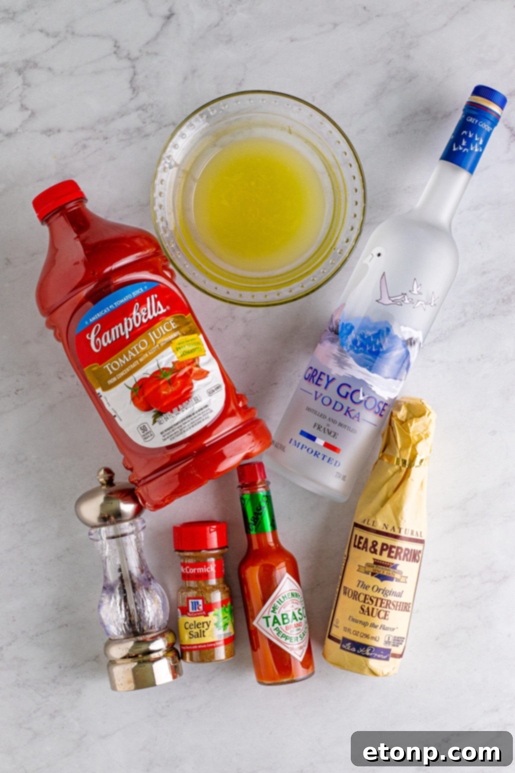 Assorted Bloody Mary drink ingredients, including tomato juice, hot sauce, Worcestershire, lemon, vodka, and spices, neatly arranged on a white marble table, ready for mixing.