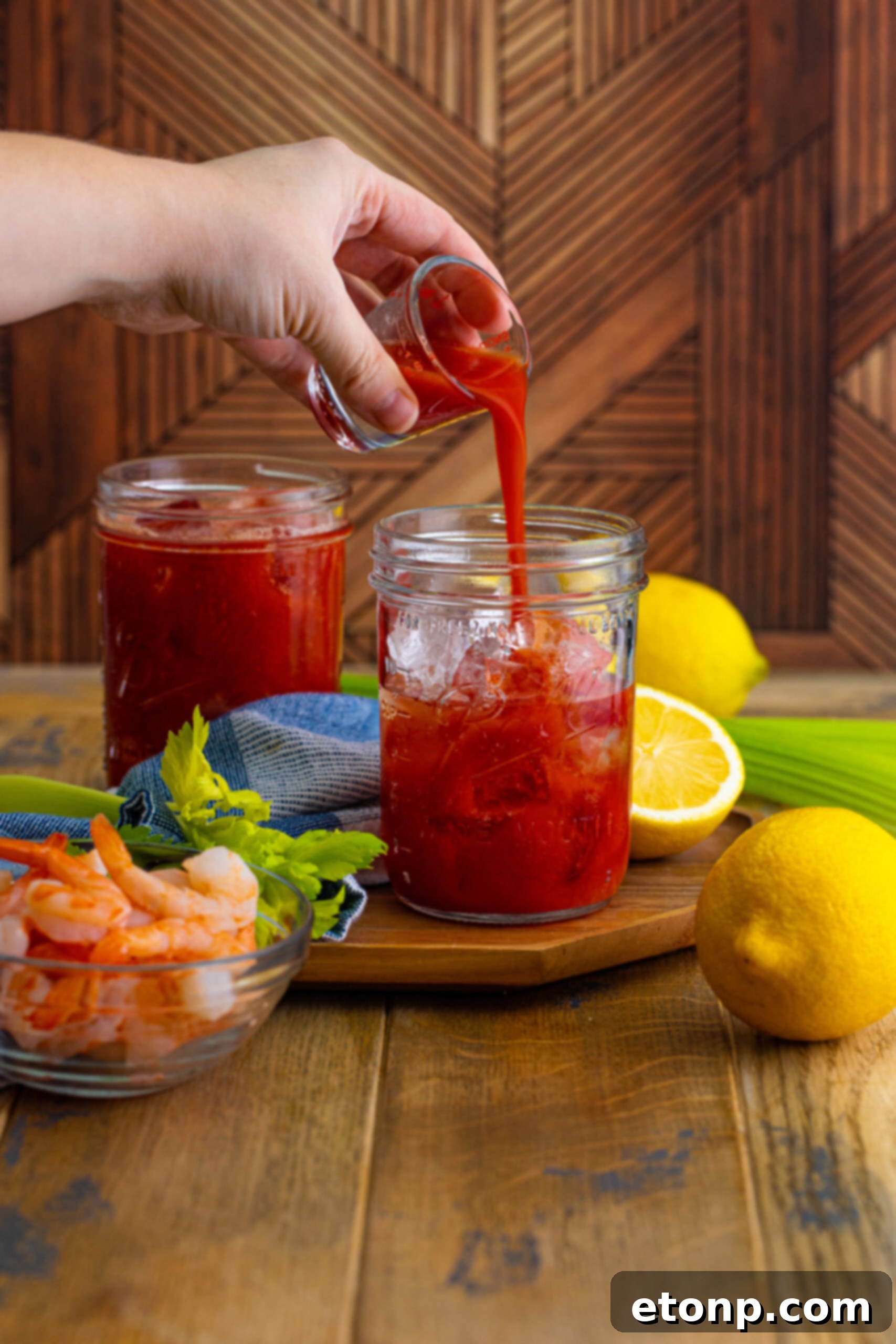 Pouring rich red tomato juice from a bottle into a glass filled with ice, preparing the base for a Bloody Mary cocktail on a rustic table.