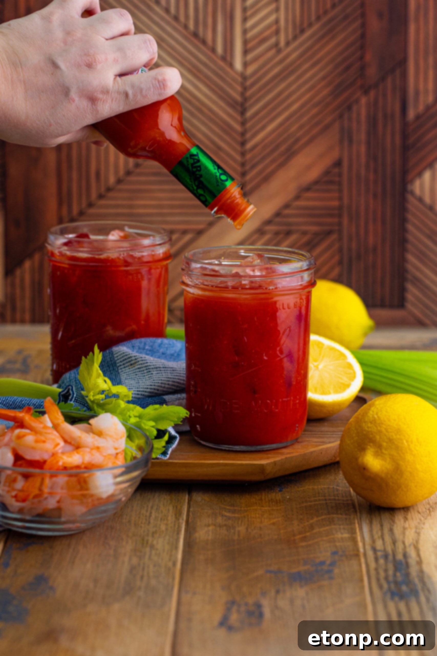 Adding dashes of hot sauce to a glass of Bloody Mary, creating the signature spicy kick, with other ingredients visible.