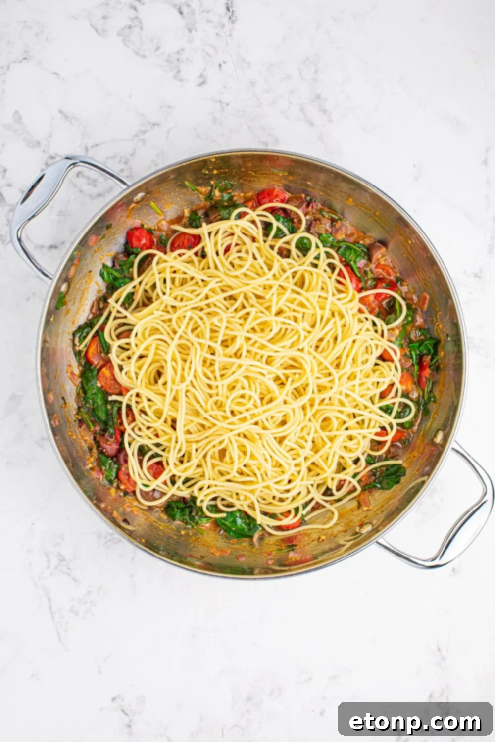 Spaghetti being tossed in a pan with the cooked vegetable mixture, ensuring every strand is coated for the Greek Spaghetti dish