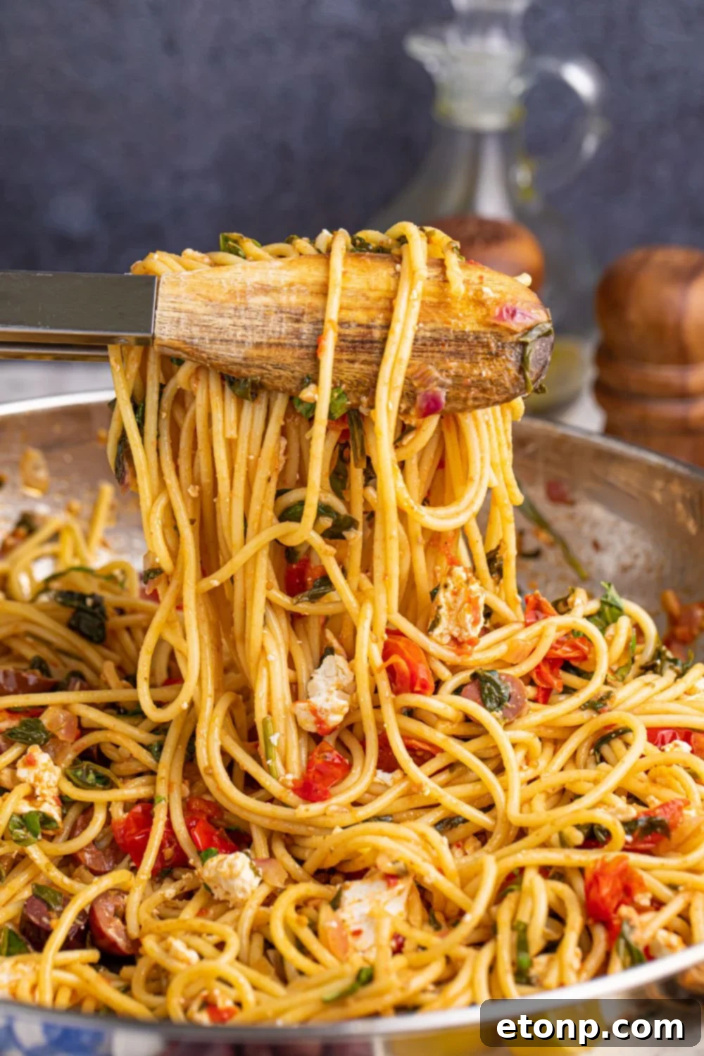Greek Spaghetti being tossed with tongs in a pan, showing the rich coating of dressing on the pasta and vegetables