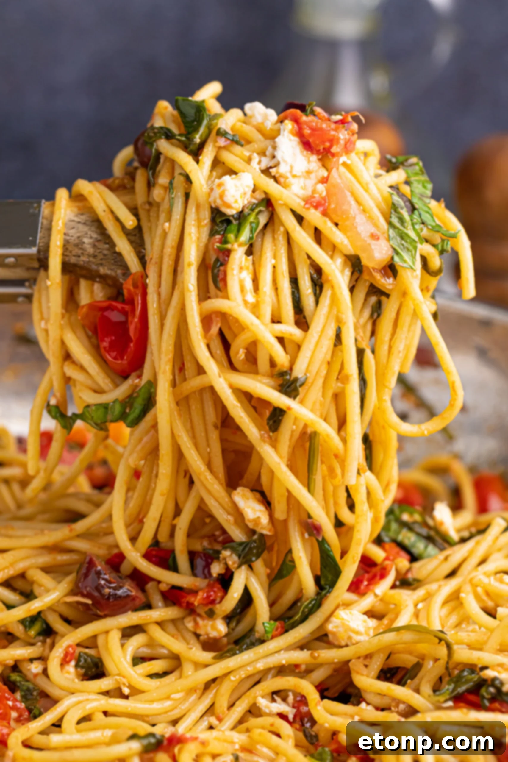 A spoon lifting a serving of Greek Spaghetti from a plate, showing the long strands of pasta intertwined with fresh vegetables