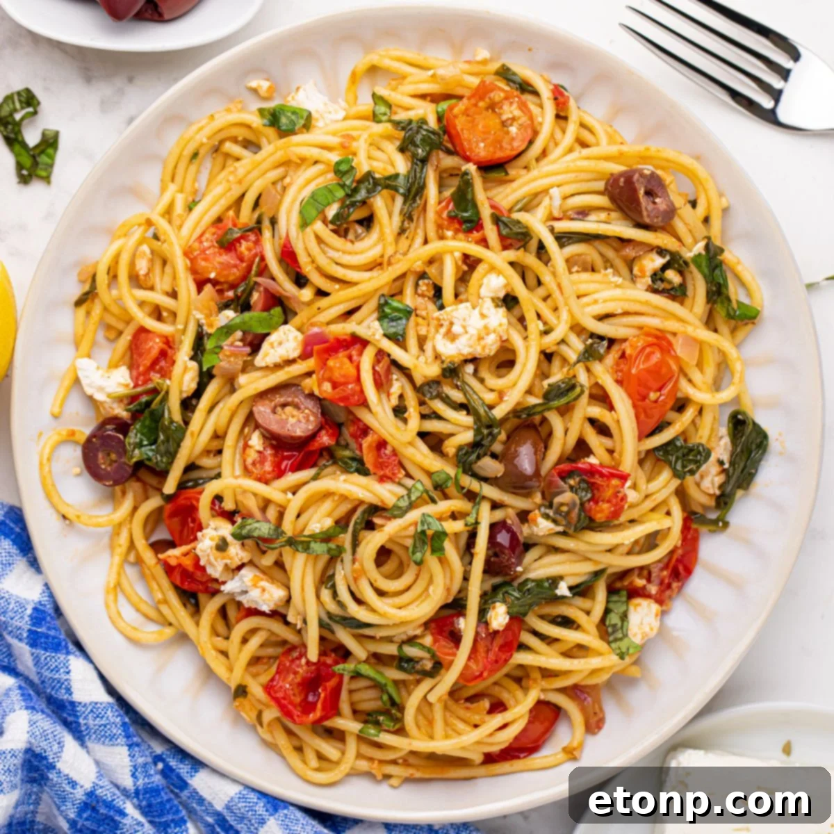 A beautifully plated Greek Spaghetti dish, with a fork resting beside it and a blue checkered towel in the background, highlighting the freshness