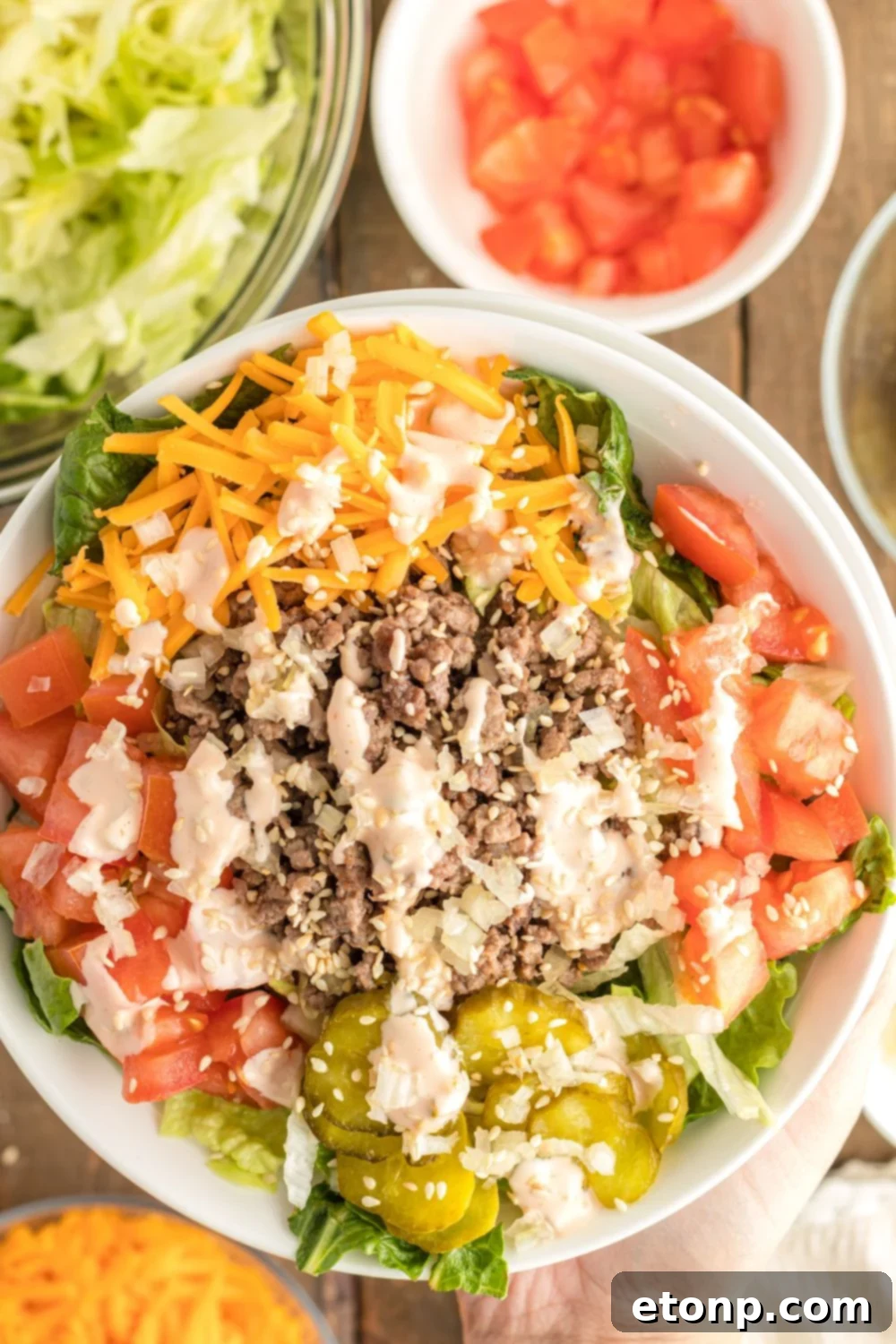 Overhead view of Big Mac Salad in a bowl, surrounded by smaller bowls containing each of its fresh ingredients: seasoned ground beef, chopped lettuce, diced tomatoes, shredded cheese, pickles, and onion.