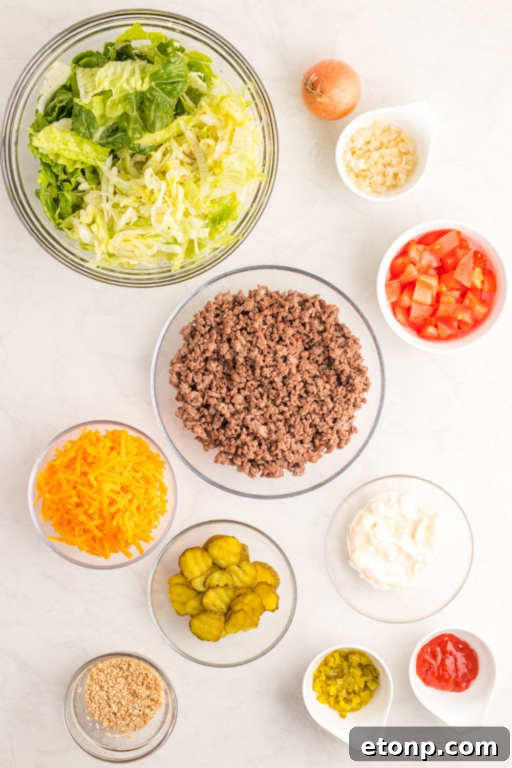 A vibrant array of Big Mac Salad ingredients neatly arranged in separate glass bowls on a white table, including fresh green lettuce, seasoned ground beef, chopped tomatoes, shredded cheese, and diced pickles.