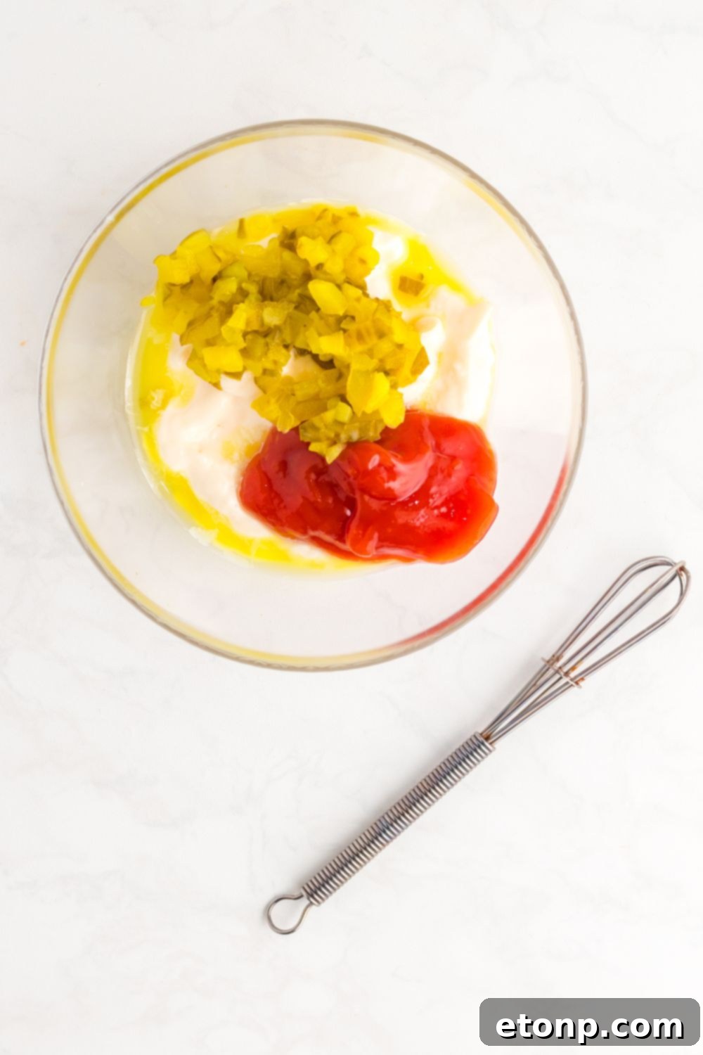 Ingredients for the Big Mac Salad secret sauce, including mayonnaise, ketchup, finely chopped pickles, and pickle juice, sit in a clear glass bowl with a whisk nearby, ready to be combined.