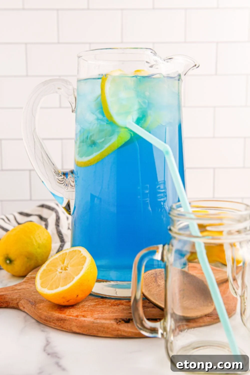 A pitcher of Blue Raspberry Lemonade on a table, with an empty glass and straw in front, inviting a refreshing pour.