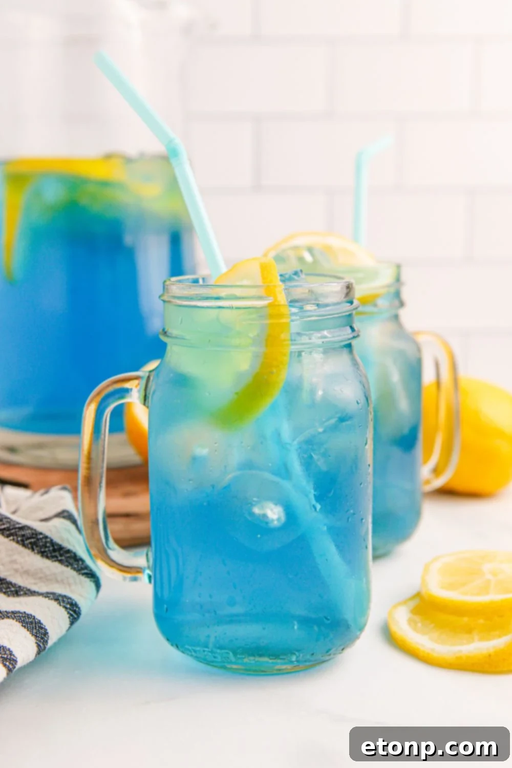 Two glasses and a pitcher of Blue Raspberry Lemonade on a table, surrounded by fresh lemon slices.