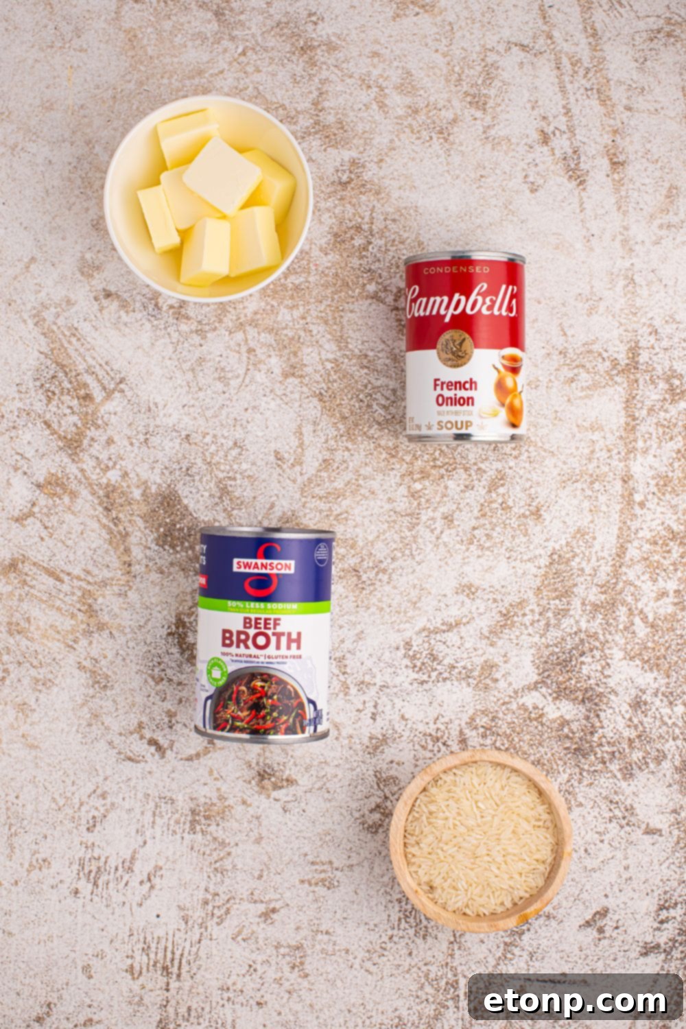 The four key ingredients for Stick of Butter Rice laid out on a table: a bowl of rice, a can of French onion soup, a can of beef broth, and a stick of butter.