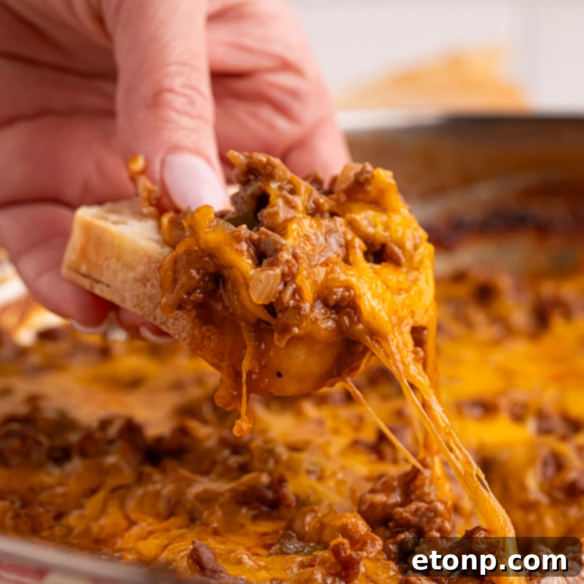 Ultimate Sloppy Joe Dip 3 A hand scooping a generous portion of cheesy Sloppy Joe Dip from a hot pan with a piece of bread, showcasing its irresistible texture.